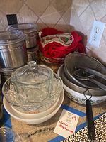 Overview of silver canisters with clear lids, Pyrex nesting mixing bowls, glass covered bowl, kitchen towels and apron, metal strainer, and fruit juicer press bowl.