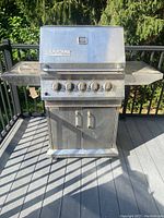 Front view of stainless steel Ducane gas grill showing five burner control knobs, side shelves, double-door cabinet, and grill cover on outdoor deck.