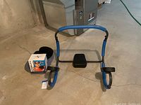 Blue ab crunch support frame with foam grips and foot pads shown on concrete floor with boxed stability ball partially visible.