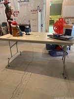 Front angled view of the folding table showing solid beige laminate top and metal legs with horizontal support bars, set up in a basement.