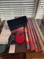 Six rolls of holiday-themed gift wrap, two small cranberry-colored beaded lamp shades, a black office desktop organizer, pack of red file folders, and a small gray pouch on a wooden table near window blinds.