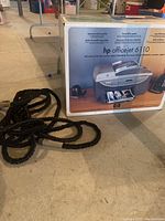 Photo of the HP OfficeJet 6110 original box and coiled black garden hose with nozzle on floor