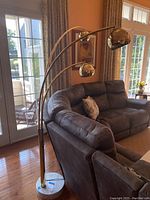 Side view of the large floor lamp placed beside a gray sectional couch showing the marble base and the full height of the lamp with three adjustable arms.