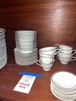 Photos show various stacks of white porcelain Noritake and Brazilian China dishes with silver rims, including dinner and salad plates, small bowls, teacups, and saucers on wooden shelves.