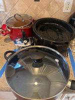 Photo of five kitchen items on a kitchen counter: red pot with glass lid, silver teapot, black roasting pan with lid, GreenPan skillet with glass lid.