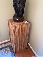 Front view of rustic wood sculpture stand with a dark carved head sculpture on top (sculpture not included in lot). The wood has natural vertical planks and textured finish.