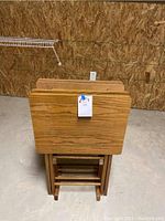 Four lightweight wooden TV tables stored upright on a wooden stand in a basement area with plywood wall.