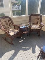 Two brown wicker rocking chairs with floral cushions and matching wicker side table with glass top on porch floor.