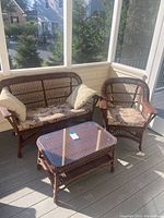 Wicker loveseat, chair, and glass top table set situated on a screened porch with sunlight casting shadows. Cushions and pillows visible on loveseat and chair.