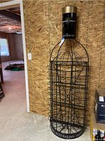 Full view of the tall metal wine rack shaped like a wine bottle against a plywood wall in a basement, showing the full height and wire slots for wine bottles.