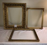Three vintage wooden picture frames displayed on white fabric, one large ornate frame and two smaller frames with decorative metal corners.