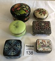 Photo of the six trinket boxes arranged closed on a white background showing their varied styles and materials such as metal, carved stone, painted floral designs, and intricate filigree work.