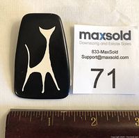 Front view of the black Hans Hansen cat brooch with white stylized cat design next to a number 71 MaxSold lot card and ruler showing size.