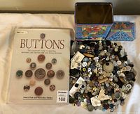 Photo showing the button reference book next to a large pile of assorted buttons and two decorative tins
