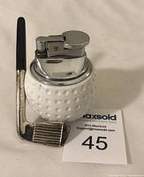 Golf-themed table lighter shaped like a white golf ball with chrome lighter top, resting next to a golf club shaped lever, showing overall front detail and shape.
