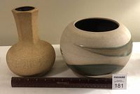 Photo showing both vases side by side with ruler for size context; left vase is textured beige with incised decoration, right vase is smooth rounded with green glaze band.