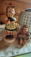 Photo of two Hummel porcelain figurines: a standing girl holding a bird and basket, and a seated boy playing an accordion.