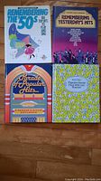 Four vintage music books laid out on wooden floor, titles visible including 'Remembering the '50s', 'Remembering Yesterday's Hits', 'Parade of Popular Hits', and 'Treasury of Favorite Songs' showing colorful covers
