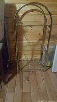 Full frontal view of three tier brass coloured metal and glass shelf with arched top, against a wooden wall background.