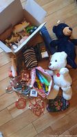 Photo showing box with plastic farm animals, zebra figurine, black and white plush teddy bears with maple leaf, bundle of colorful markers in pouch, and small toy trucks arranged on wooden floor.