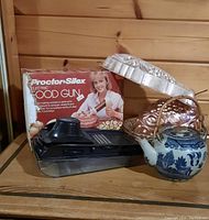 Various vintage kitchen items on wooden shelf including the Proctor Silex electric food gun box, mandolin slicer, Chinese tea pot, and jelly moulds.