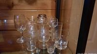 Photo showing 12 assorted clear glasses including wine glasses, snifters, tumbler, and shot glass on wooden surface.