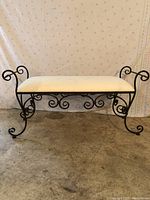 Front view of wrought iron bench with beige upholstered seat and decorative scrollwork on legs and sides.
