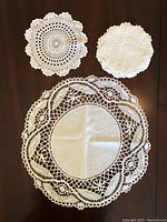 Photo shows three round linen doilies including one large handmade antique Irish linen piece (17 inches) with detailed lace lace edging, one medium 9-inch round crocheted doily, and a smaller white crocheted doily, all laid on a dark wooden surface.