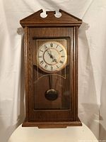 Full frontal view of the wooden miniature grandfather wall clock with glass door and visible brass pendulum.