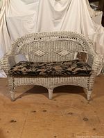 Front view of white painted wicker loveseat with floral cushion, showing woven diamond pattern on backrest and six legs.