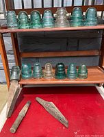 Photo of two shelves holding 11 glass and porcelain insulators in various shapes and green to clear colors, with two wooden pegs on red surface below.