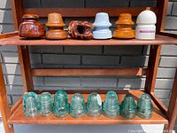 Photo of collection of glass and porcelain electrical insulators arranged on wooden shelves outdoors, showing variety in shapes and colors.