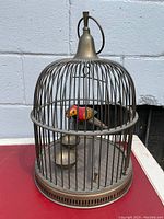 Full view of the solid brass dome bird cage showing the mechanical painted bird, perch, and bowls inside against an outdoor backdrop.
