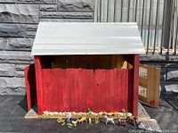 Frontal view of the red wooden barn doll house with white roof and farm animal figures arranged in front on a black surface against a stone wall background.