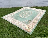 Full view of large green and white wool rug laid flat on grass showing floral central medallion and white floral border.