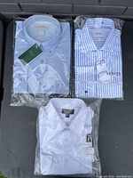 Three men's dress shirts still in packaging, colors blue solid, blue/white stripes, and white floral pattern.