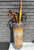 Front view of the oriental ceramic umbrella stand containing multiple wooden walking sticks and umbrellas, standing against a gray brick wall.