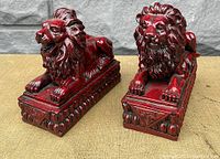 Pair of red resin Chinese lion statues resting on rectangular bases with carved geometric detail.