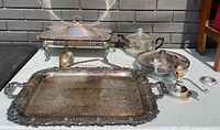 Wide angled photo of all silver plated serving pieces including tray, chafing dish, glass bowl, candlesticks, and ladle, showing condition and details.