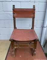 Front view of vintage Spanish Colonial Revival style chair showing leather seat and backrest with decorative metal studs and carved wooden frame.