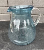 Teal bubble art glass pitcher with clear handle shown from front angle against brick background on black surface.