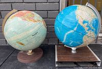 Two vintage globes side by side showing different map styles and bases on a dark surface against a brick background.
