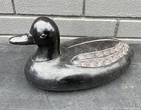 Side view of the black painted wooden mallard duck decoy on a gray surface against a brick wall, showing the carved wing feather details and rounded body shape.