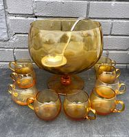Full set of amber glass punch bowl with ladle inside and 10 cups arranged around it on gray surface against brick wall.