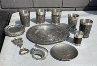 All vintage pewter serving pieces arranged on a table, showing cups, plate, small dish, coasters, bottle opener, and napkin ring.