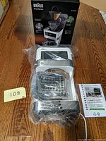 Photo of Braun BrewSense coffee maker in plastic wrap with box behind it on wooden floor, includes instruction leaflet.