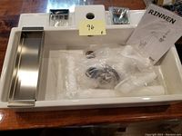 Top view of white Ikea Lillangen bathroom sink with stainless steel draining racks, miscellaneous white plumbing parts in plastic bag, metal mounting hardware, and Rinnen instruction manual arranged inside the sink basin.