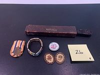 Top view of the entire lot showing the military medal with ribbon, mosaic bracelet, small ceramic plate, two miniature embroidered pieces, Allegro sharpener box, and price note.