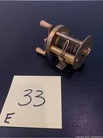 Brass fishing reel showing dual handles and the spool with thread wrapped around it, placed on a dark surface.