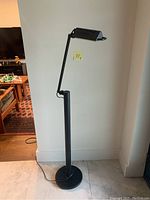Photo of black cast iron floor lamp standing against wall, showing lamp in off state with visible cord on floor.